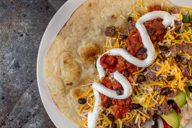 open taco tortilla with tomatoes, sour cream, bean, and beef with cheese flat lay
